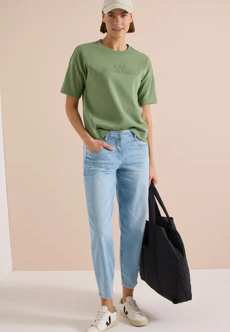 Frau trägt grünes T-Shirt, helle Jeans und hält schwarze Tasche
